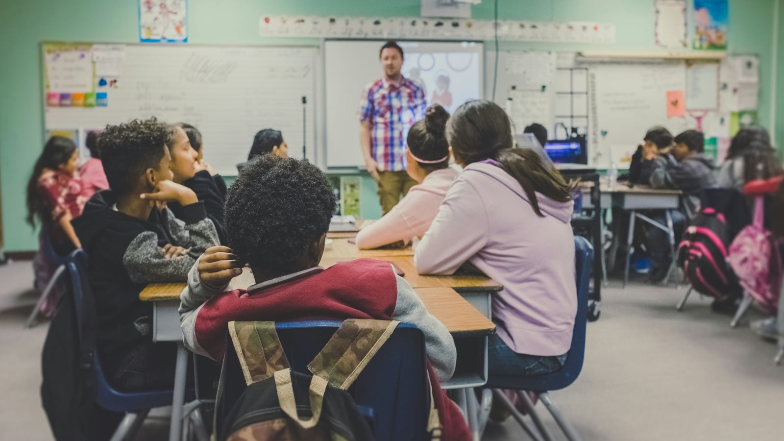 Students at their desks in a classroom, listening to a teacher standing at the whiteboard